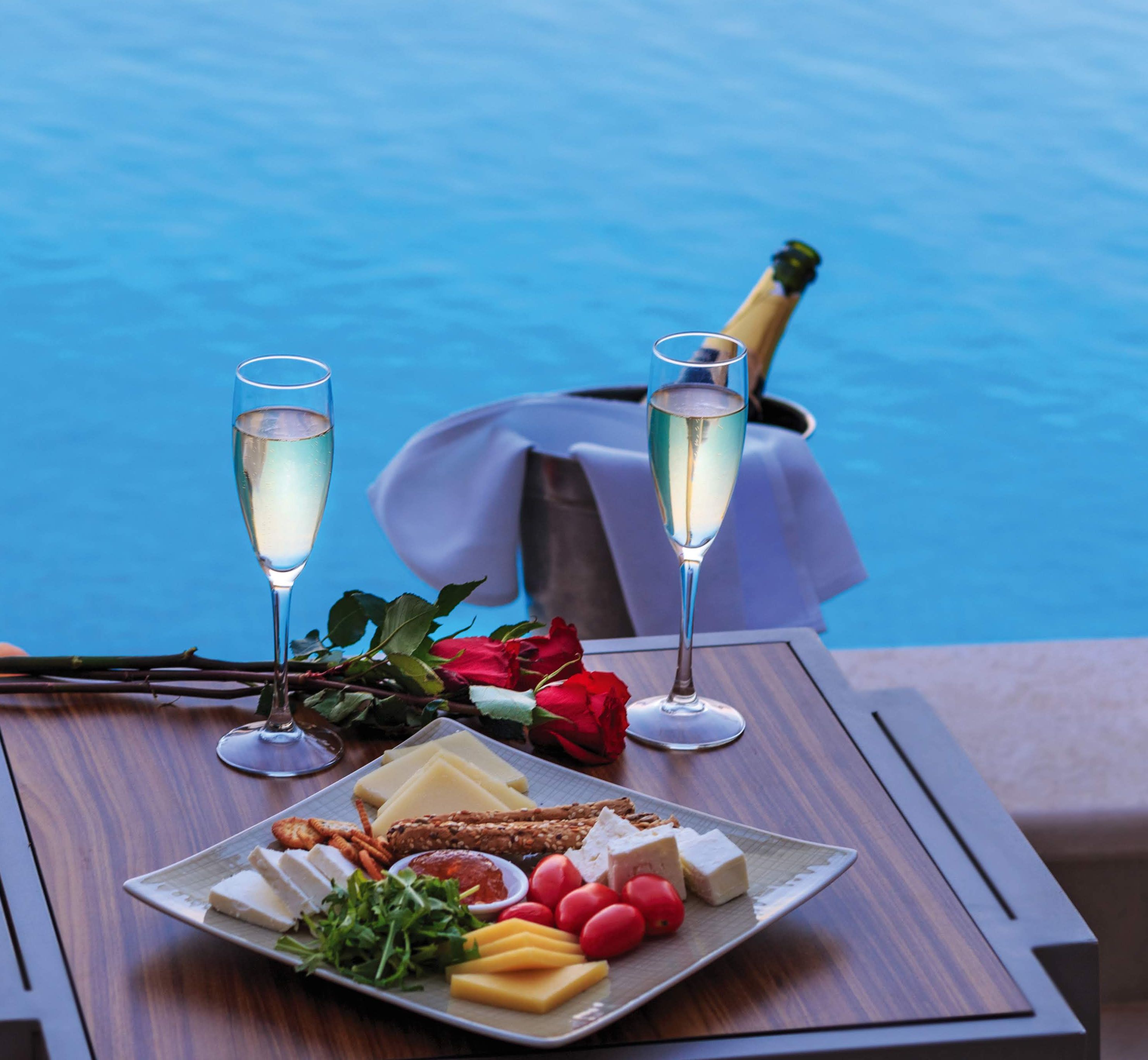 A table with a plate of cheese, crackers, and a bouquet of red roses, two champagne flutes, and a bottle of champagne.