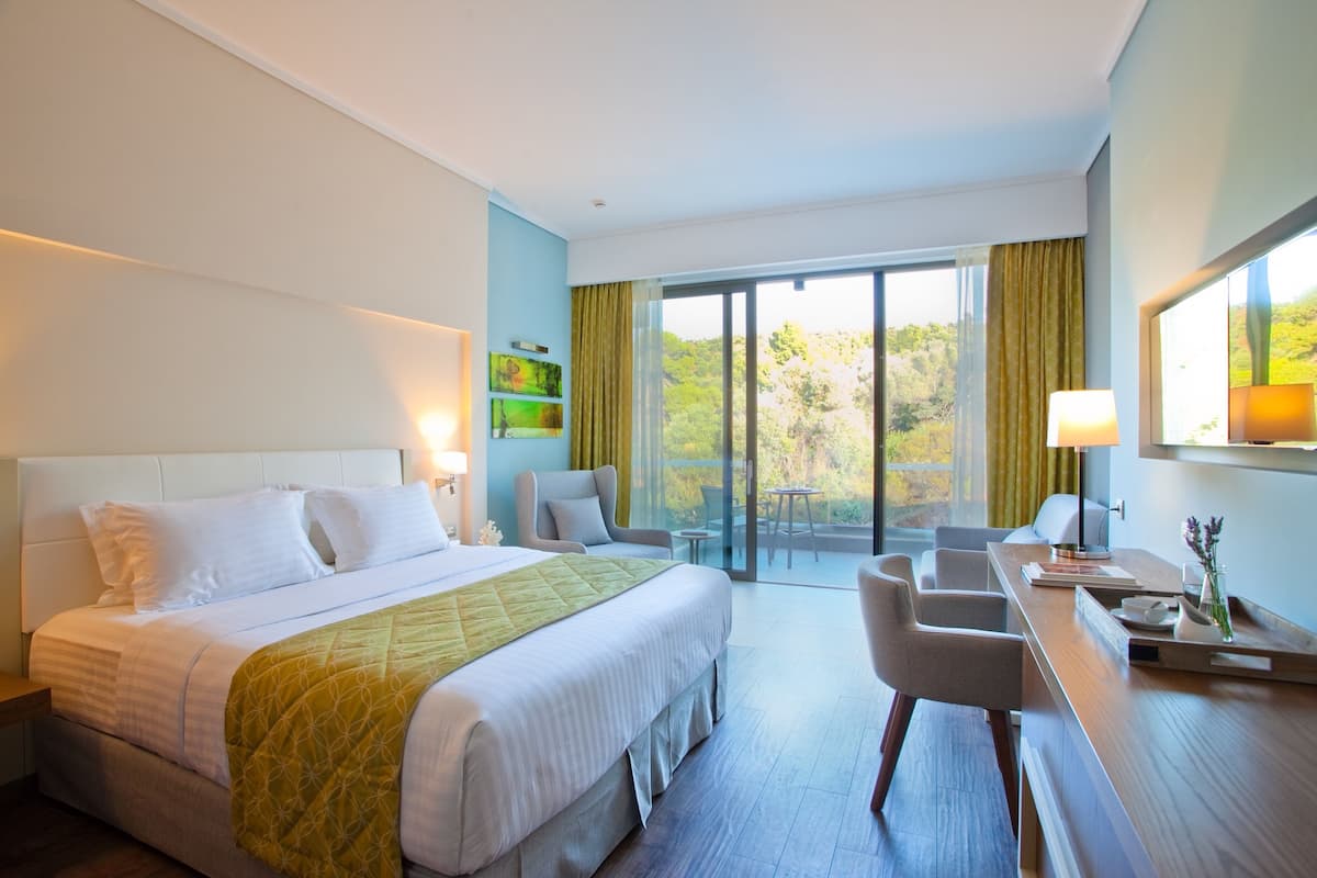 Ein Hotelzimmer mit einem großen Bett, einem Schreibtisch mit Stuhl, einem kleinen Tisch und Blick ins Freie durch eine Glastür.