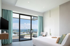 Habitación de hotel moderna con una cama grande, televisor de pantalla plana, escritorio y vistas al océano y al puerto a través de ventanas de techo a piso.