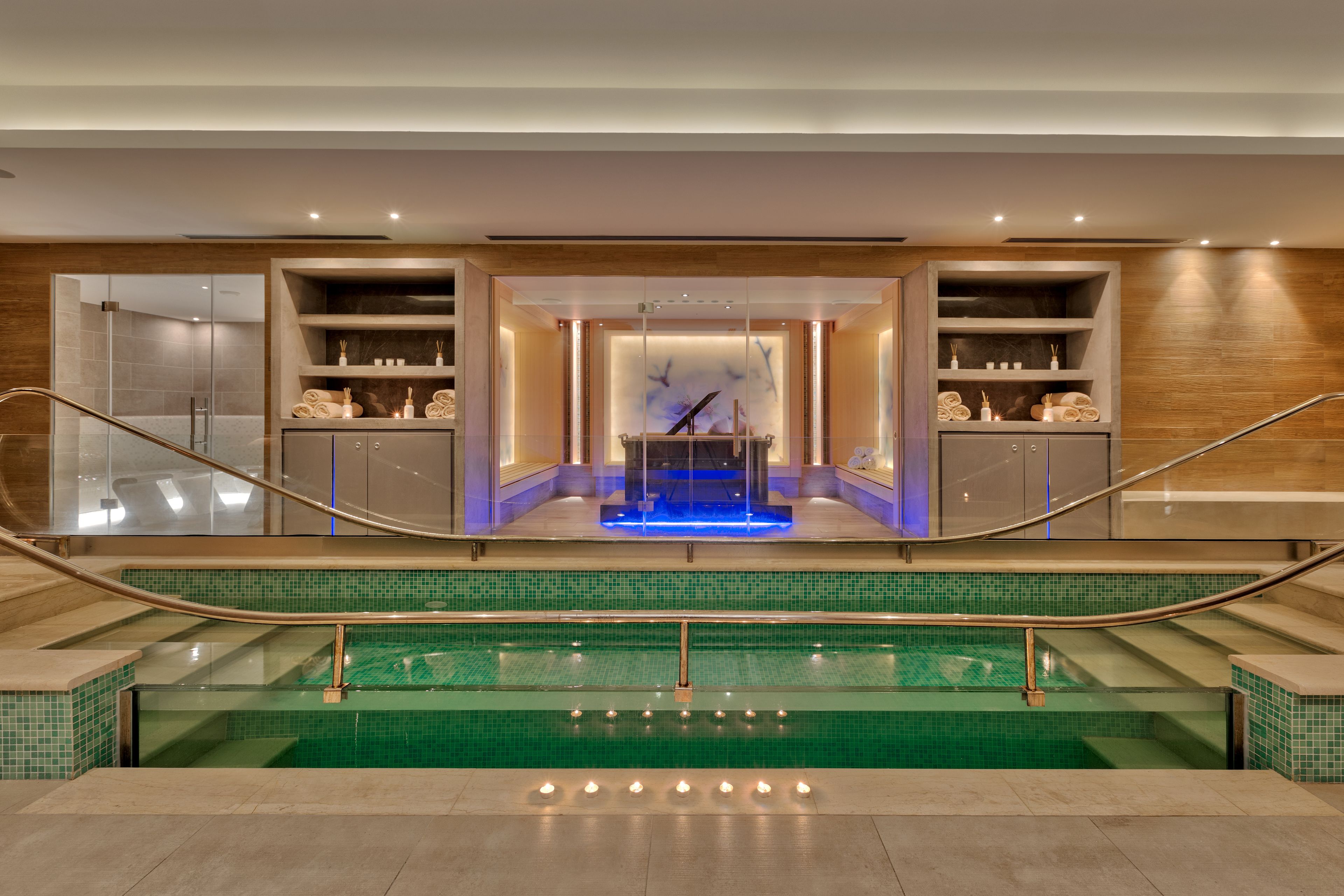 Un espace piscine intérieur moderne, doté d’une paroi en verre, d’un élément décoratif aquatique illuminé de lumière bleue, ainsi que d’étagères contenant des serviettes et des articles de toilette.