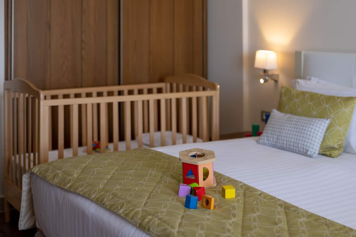Ein ordentlich bezogenes Bett mit einem gemusterten Überwurf, ein Holckrippe im Hintergrund und ein Nachttisch mit Lampe und Kissen.