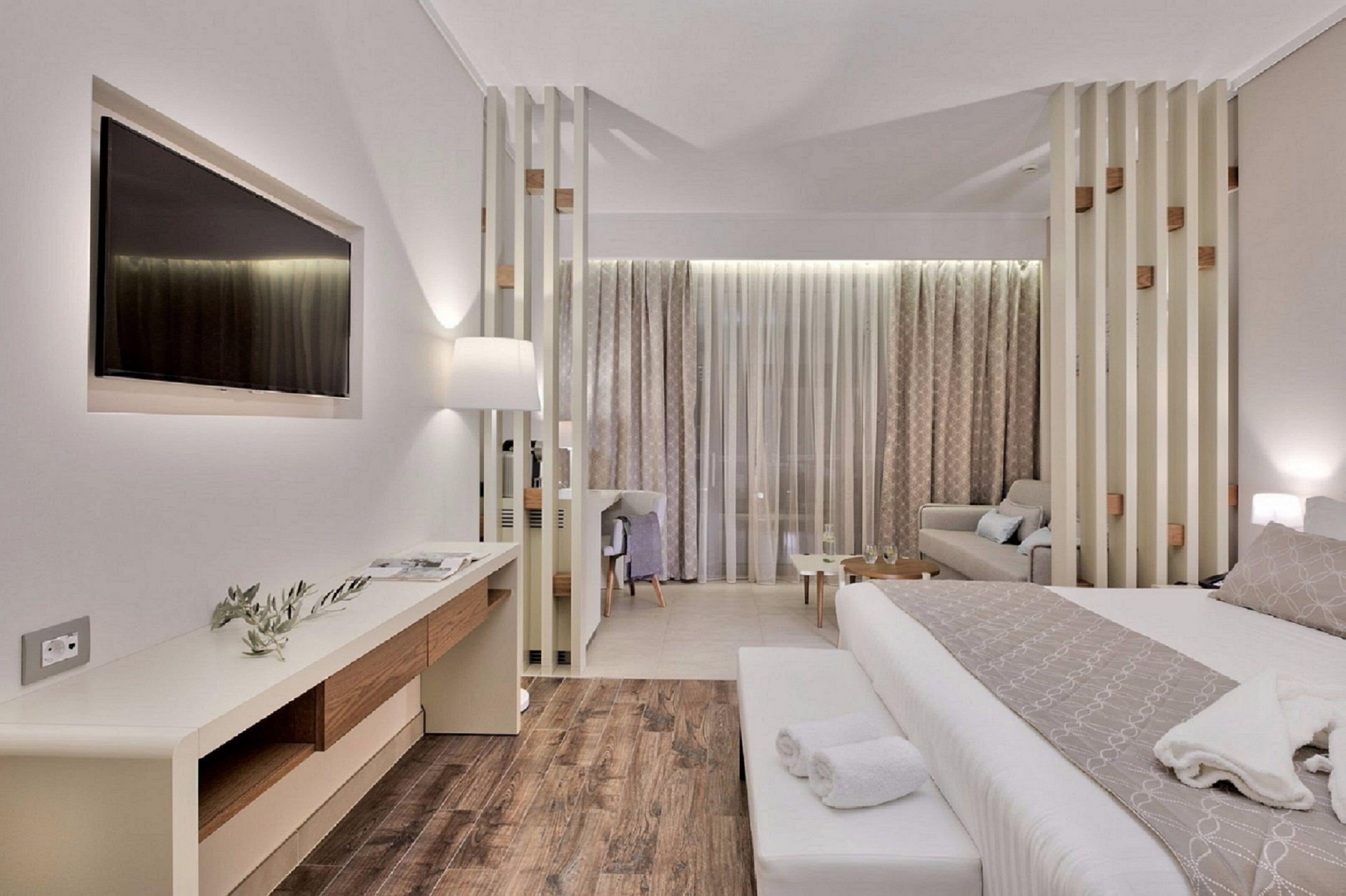 A modern hotel room with a large bed, a bedside table, a desk with a chair, a TV, and a large window with curtains.