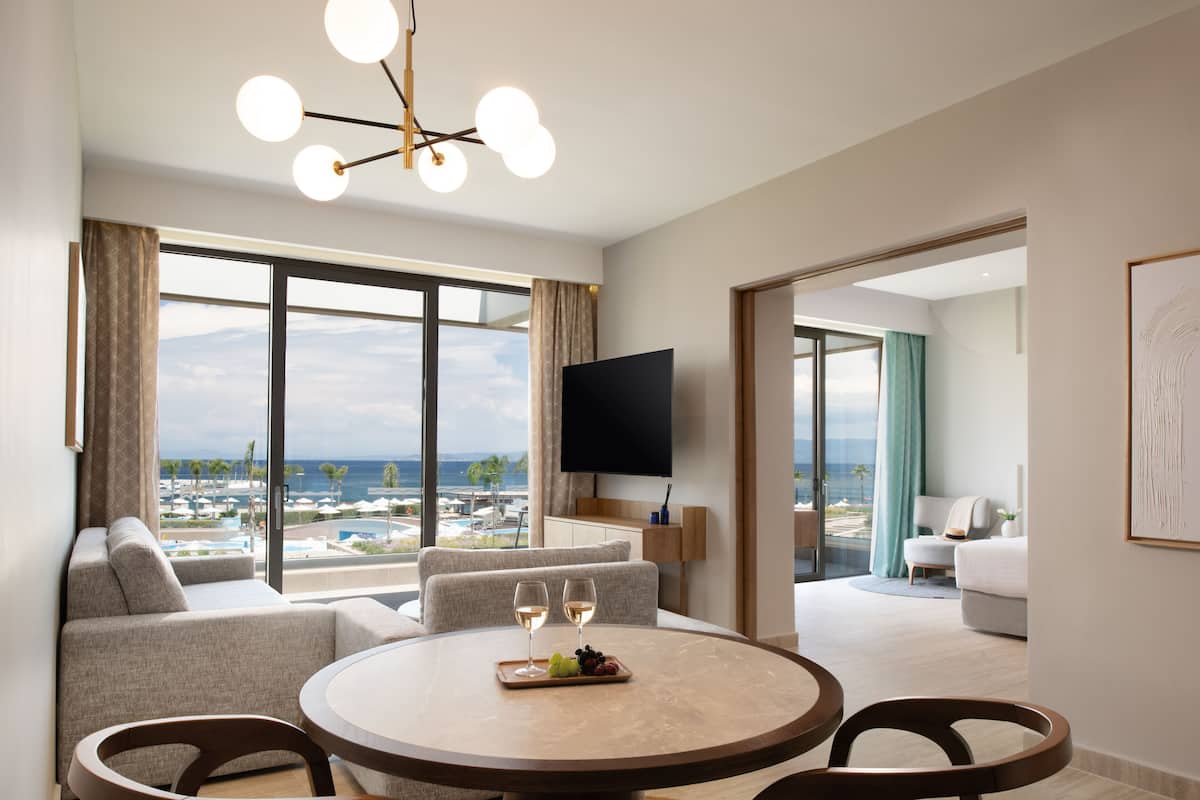 Ein modernes Hotelzimmer mit großem Fenster und Blick auf Strand und Meer. Es gibt einen runden Esstisch mit zwei Weingläsern und einer Obstplatte.