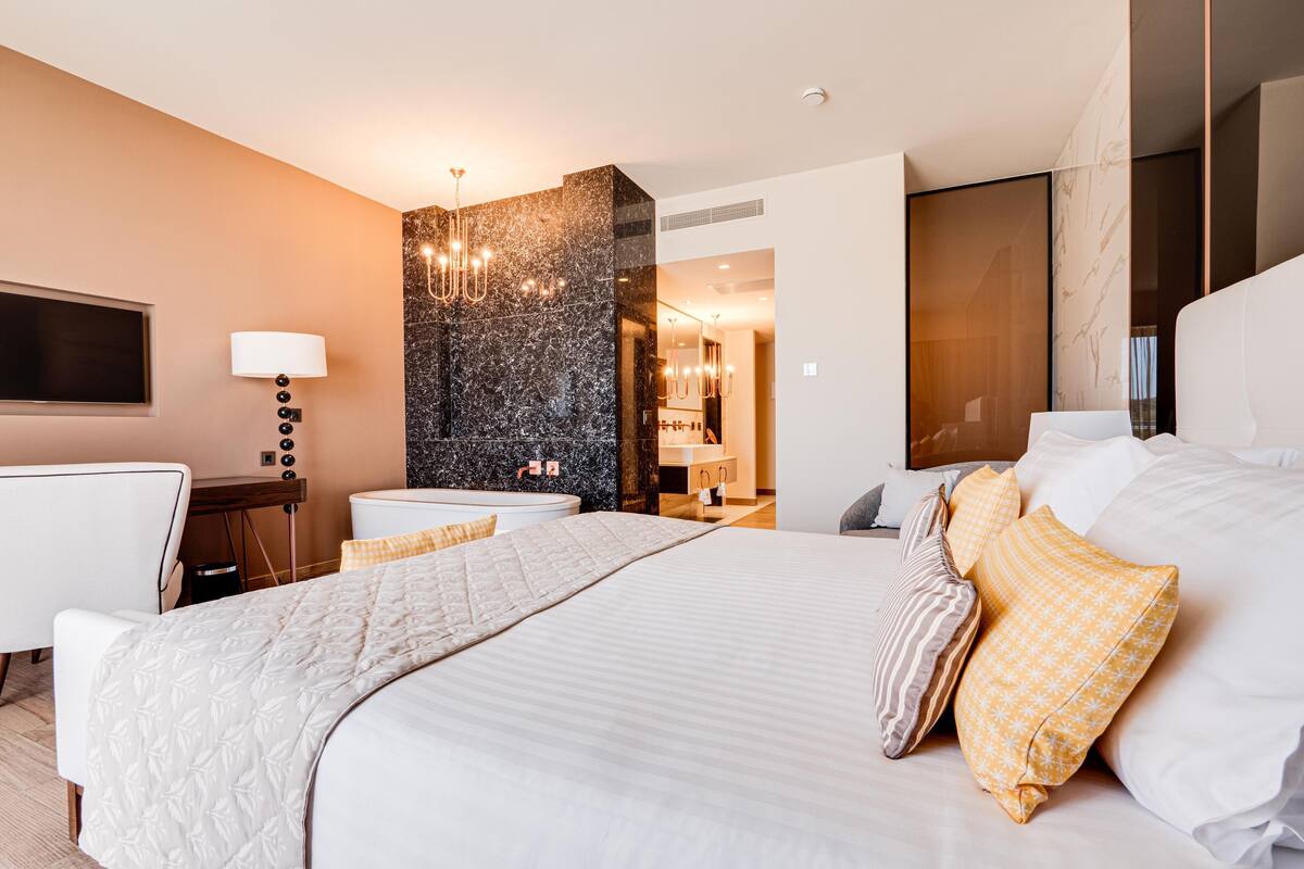 Ein modernes Hotelzimmer mit einem großen Bett, einem Schreibtisch, einem Sessel, einem Fernseher und einem durch eine offene Tür einsehbaren Badezimmer.
