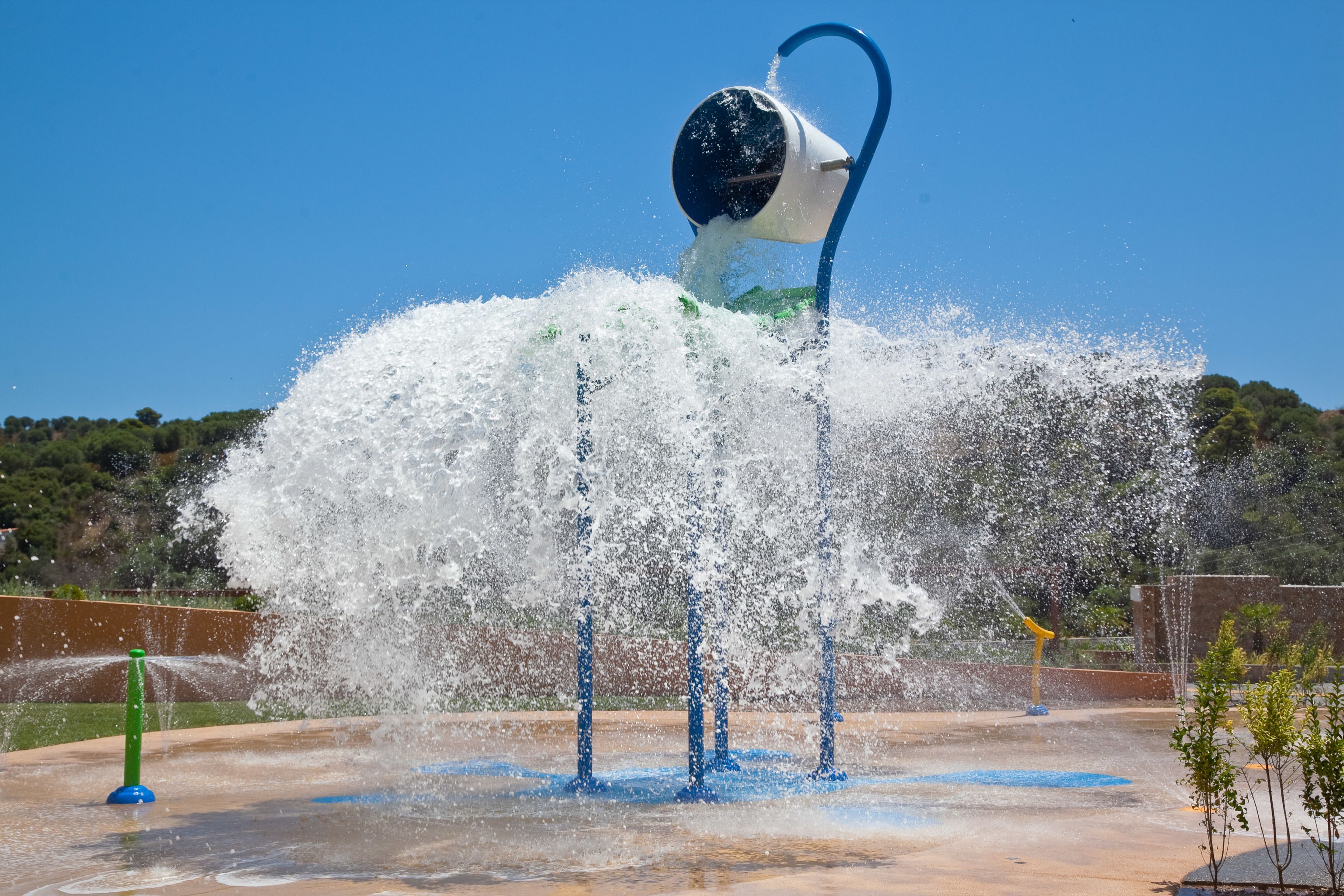 A water fountain with a blue and white structure, surrounded by a clear blue sky and greenery.