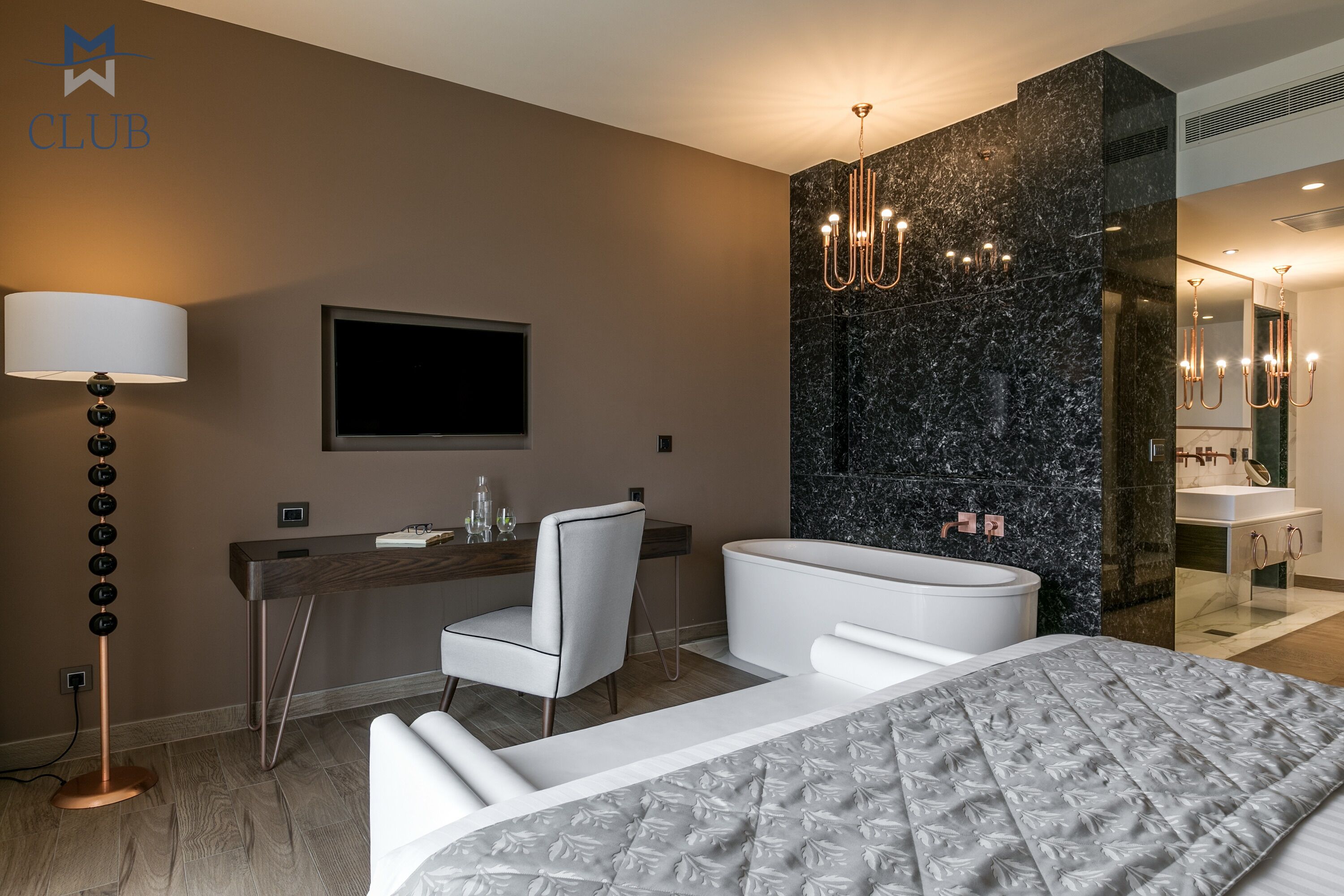A modern hotel room with a bed, a desk, a chair, a TV, a chandelier, and a bathroom.