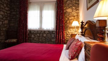 A room with a stone wall, a bed with red bedding, a nightstand with a lamp, and a window with curtains.