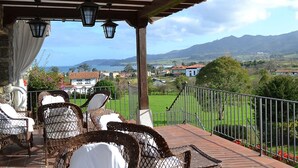 A covered outdoor seating area with wicker chairs and a view of a green landscape and mountains in the distance. - Hotel Los Caspios (Colunga)