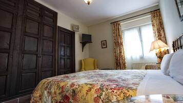 Un dormitorio con una cama grande, una silla, un televisor en un estante y una ventana con cortinas.