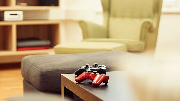 A gaming controller on a wooden table in a living room with a green armchair and a shelf with books and a TV.