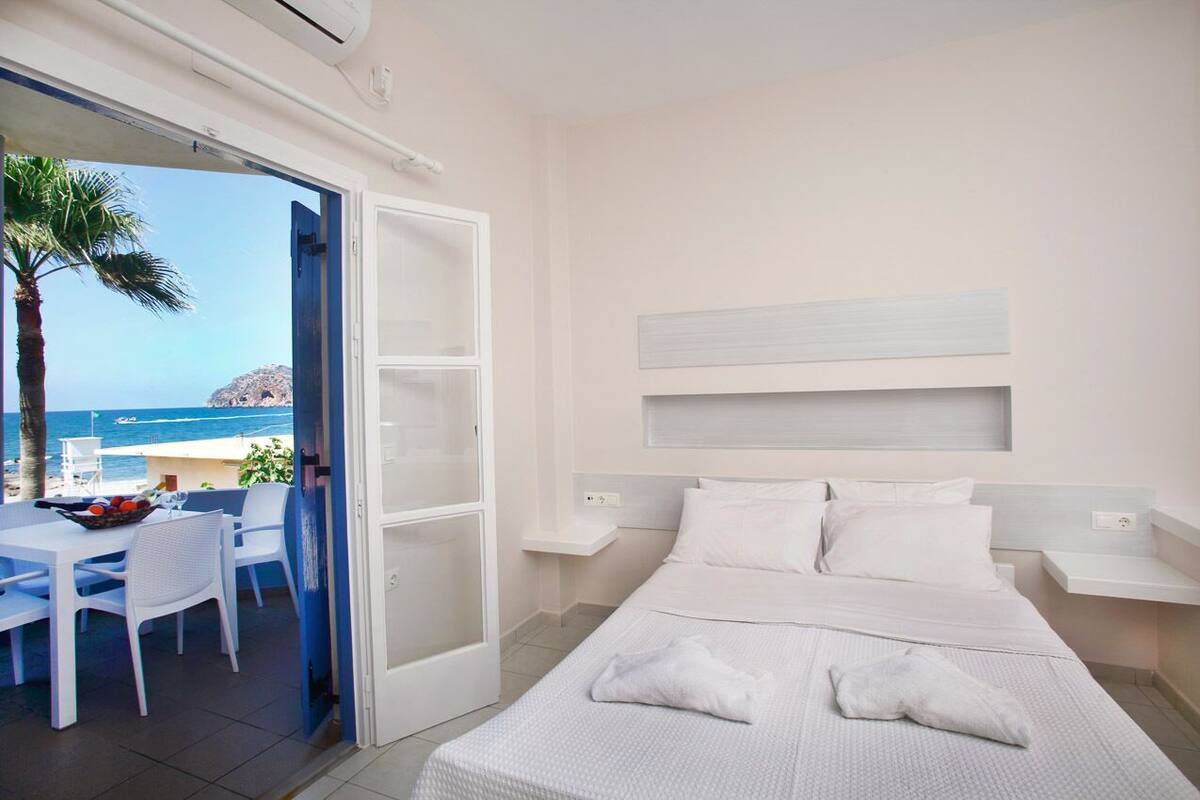 A hotel room with a bed, a small table with chairs, a view of the sea, and a palm tree outside.