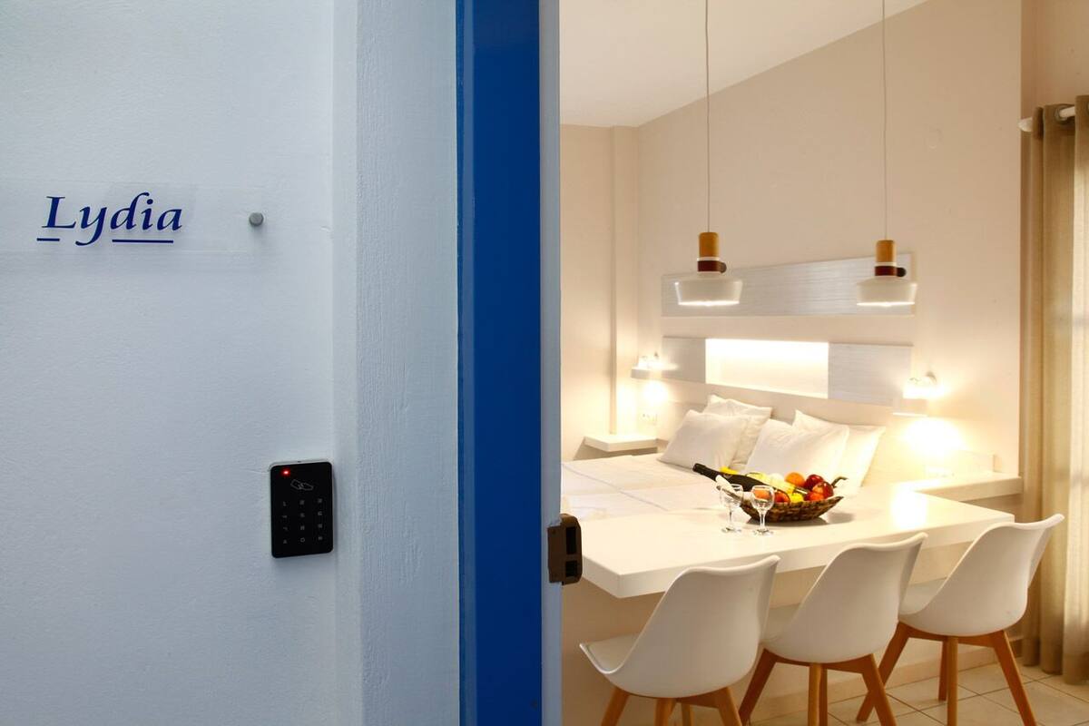 A modern hotel room with a white wall, a blue accent wall, a digital lock, a white dining table with four chairs, and a fruit bowl on the table.