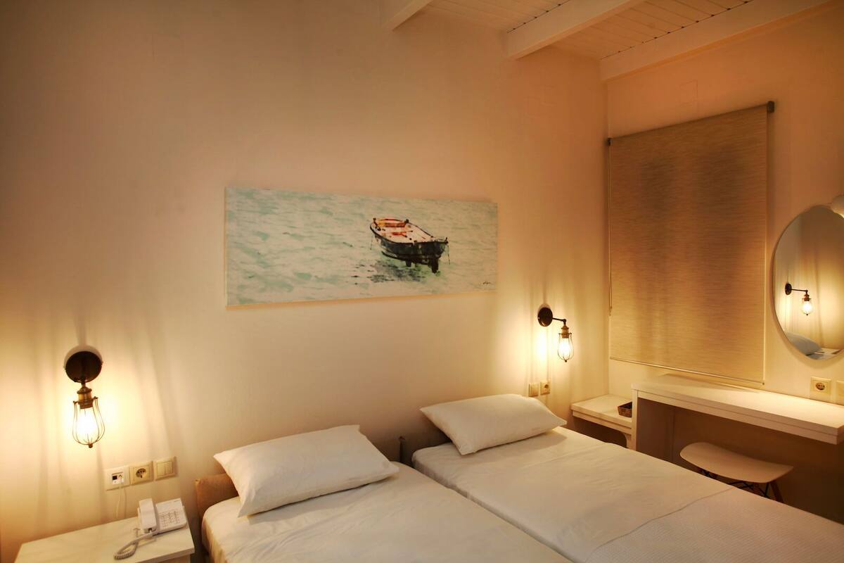 A hotel room with two beds, a painting of a boat on the wall, a bedside table with a lamp, and a mirror above the table.