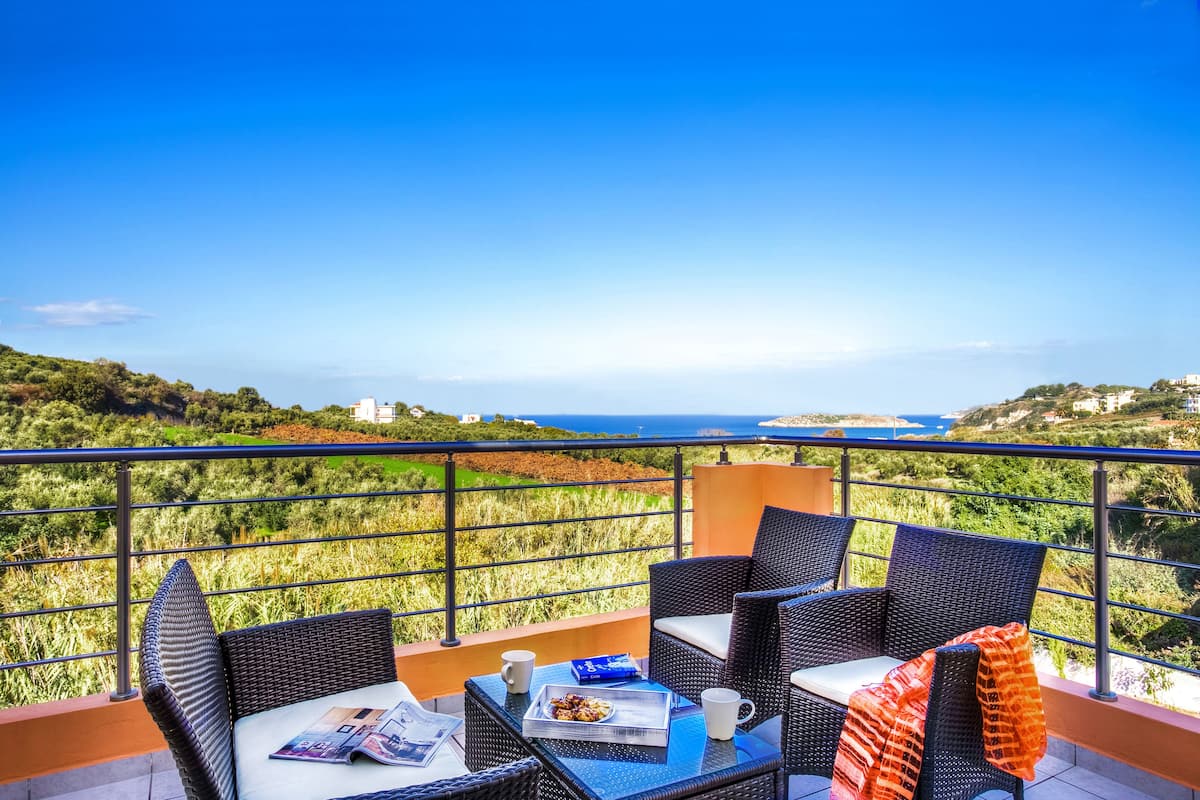 A balcony with wicker furniture, a table, and a view of the sea and greenery.