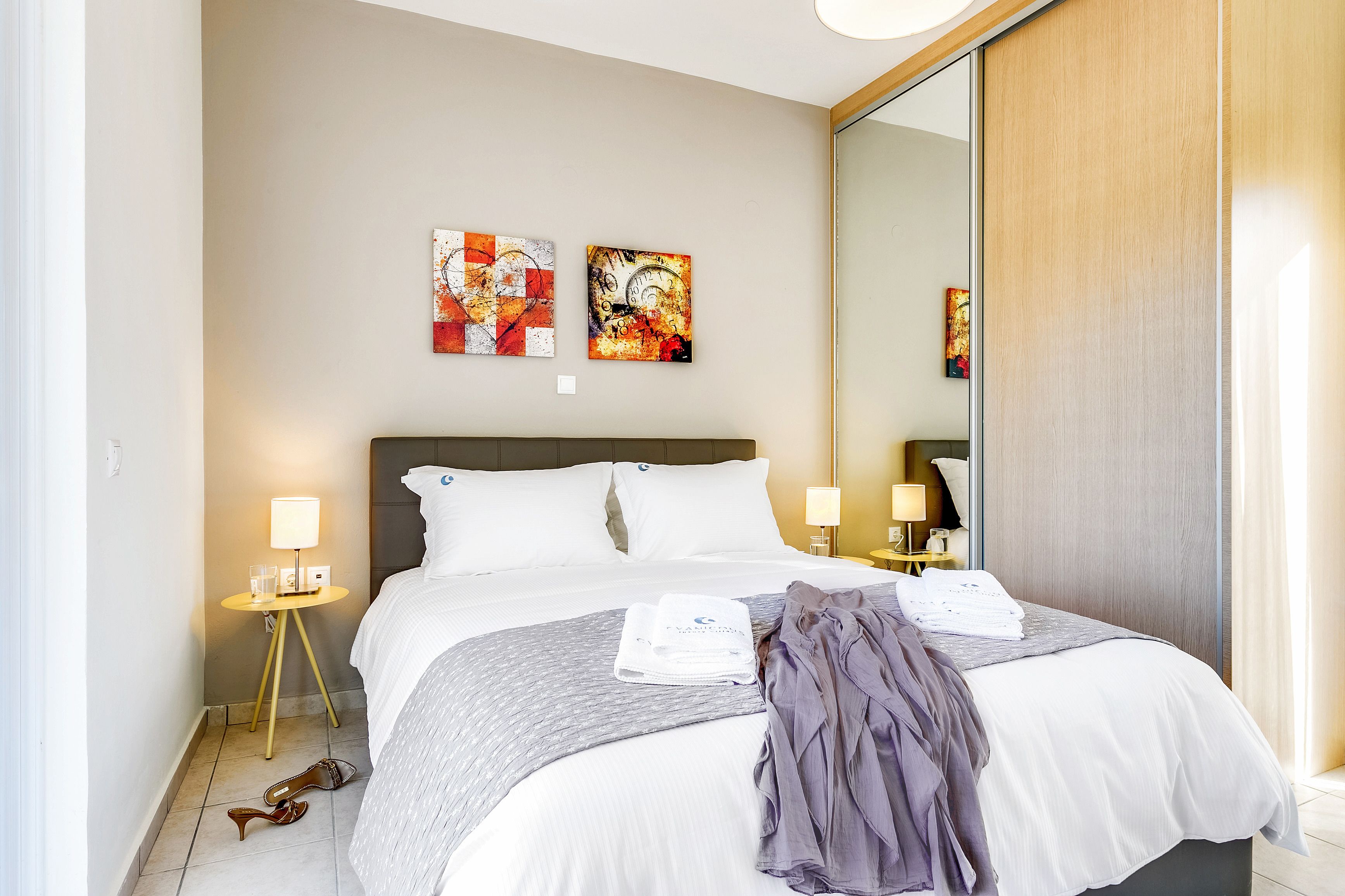Cama bem arrumada com lençóis brancos, manta cinza e um cobertor roxo. Duas pinturas na parede, um abajur em uma mesinha de cabeceira e um espelho na parede.