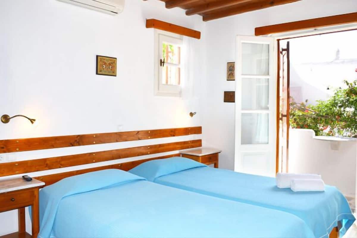 Ein Hotelzimmer mit einem hölzernen Bett, einem Nachttisch, einem kleinen Fenster und Blick ins Freie.