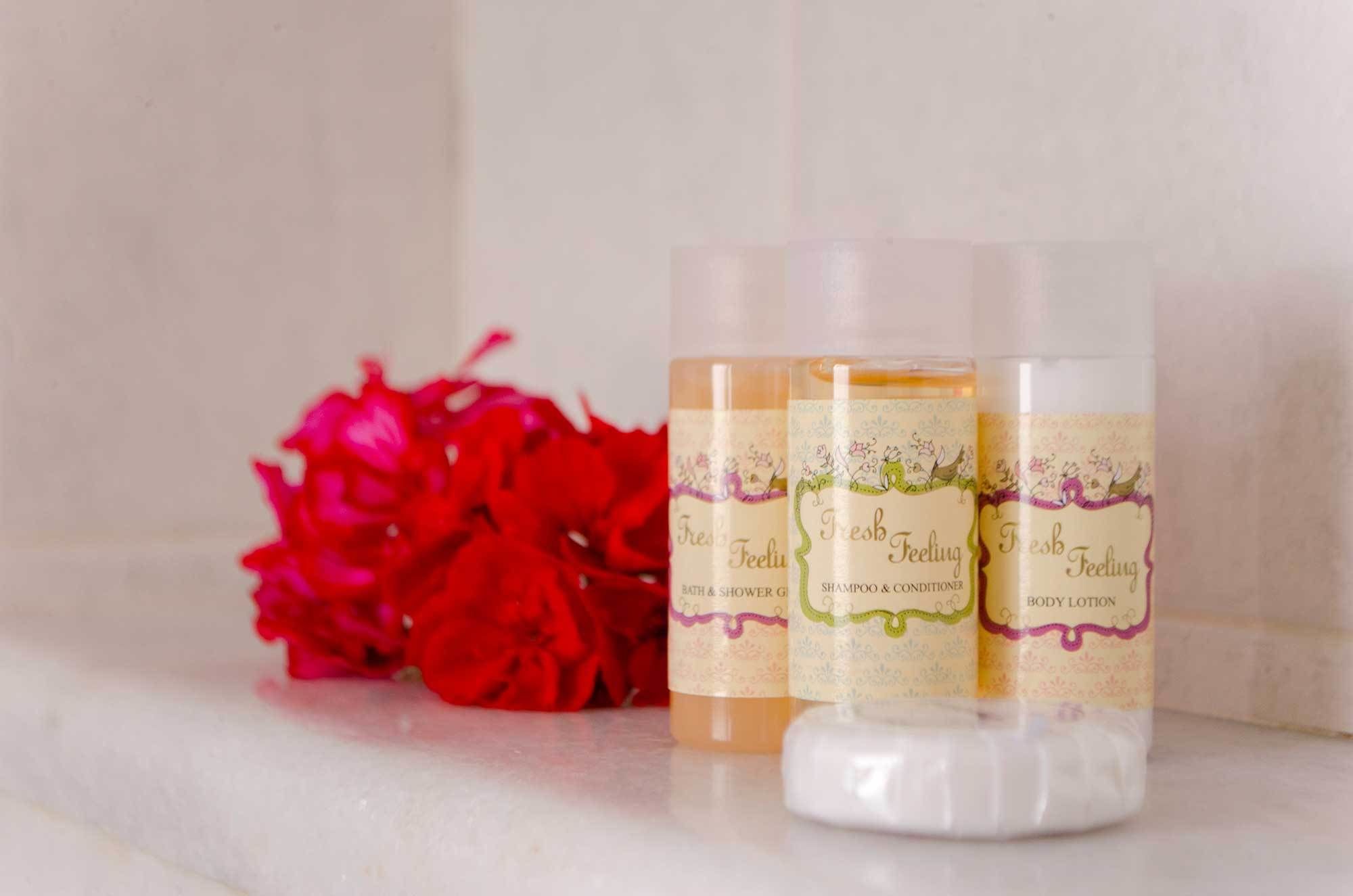 Three bottles of 'Fresh Feeling' bath and shower gel, shampoo, and conditioner, and body lotion on a white surface with red flowers in the background.