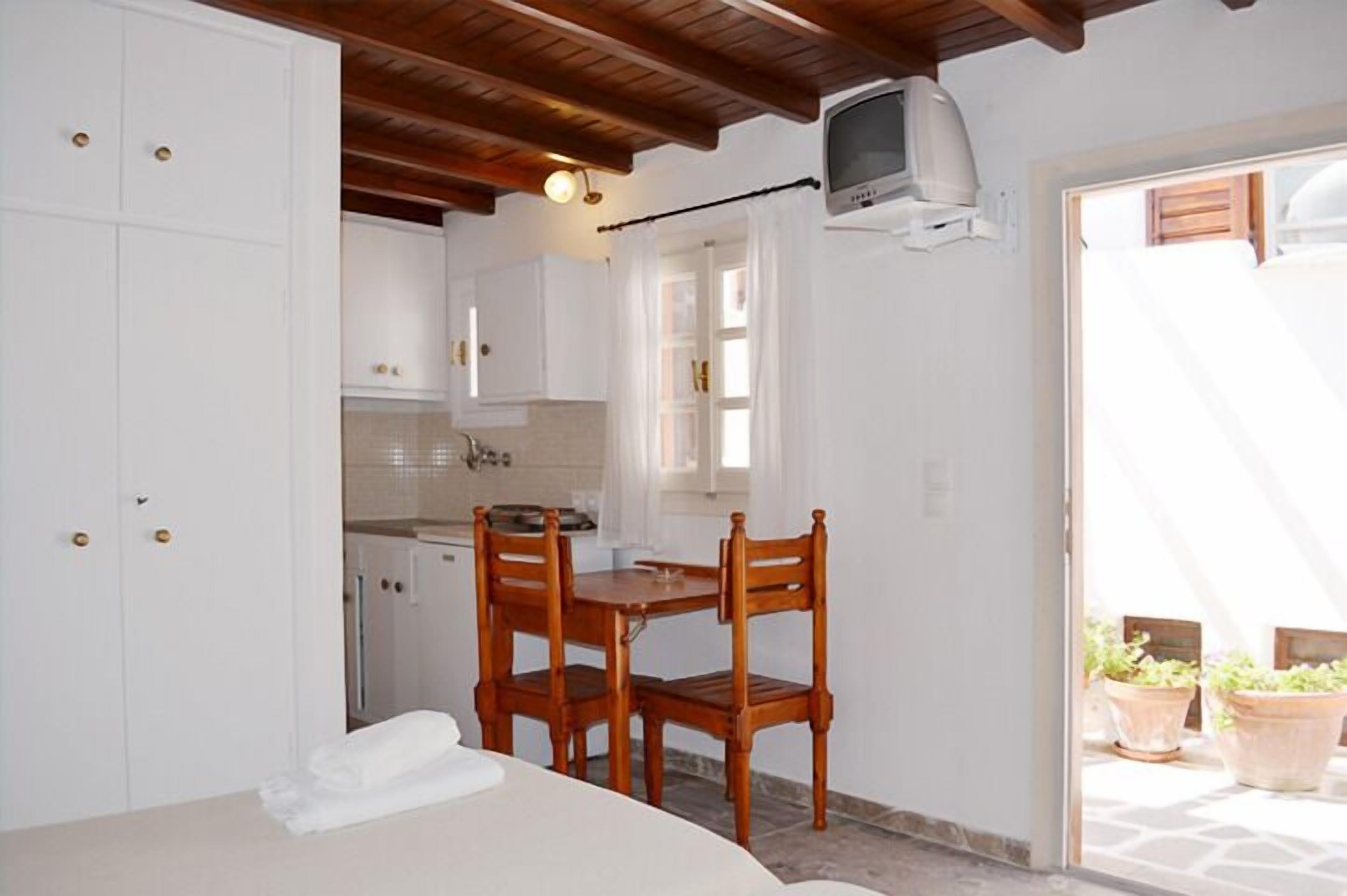 A compact kitchen with white cabinets, a wooden dining table with two chairs, and a small television mounted on the wall.