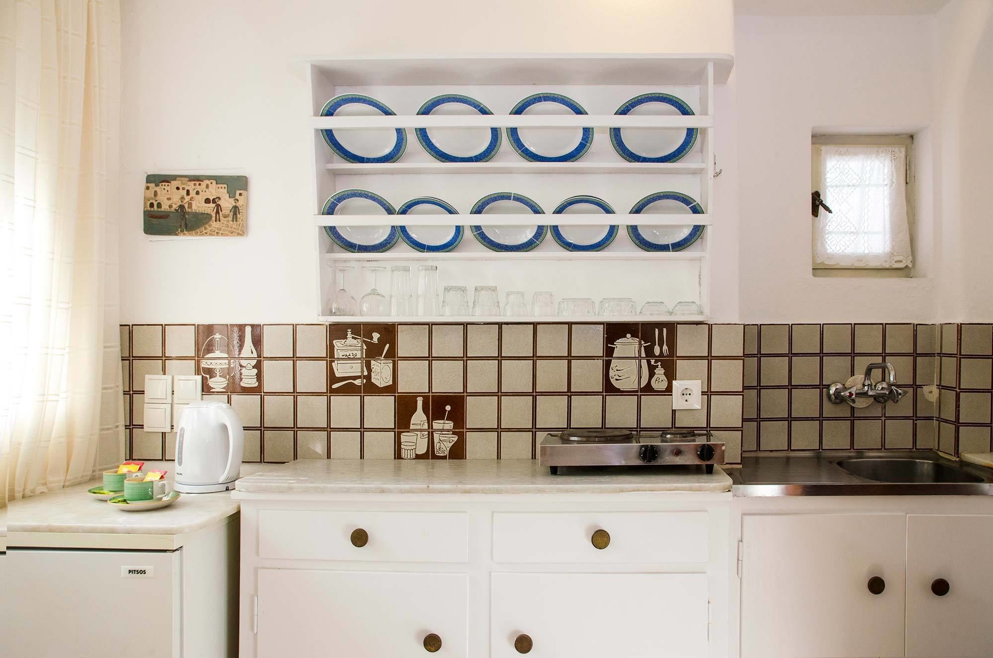 Une cuisine avec des armoires blanches, une cuisinière encastrée et un évier. On peut voir des assiettes décoratives accrochées au mur et une petite fenêtre avec un rideau.