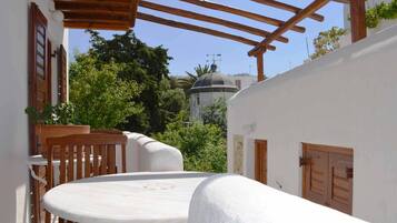A covered outdoor seating area with a round table and wooden chairs, overlooking a white building with a dome and lush greenery.