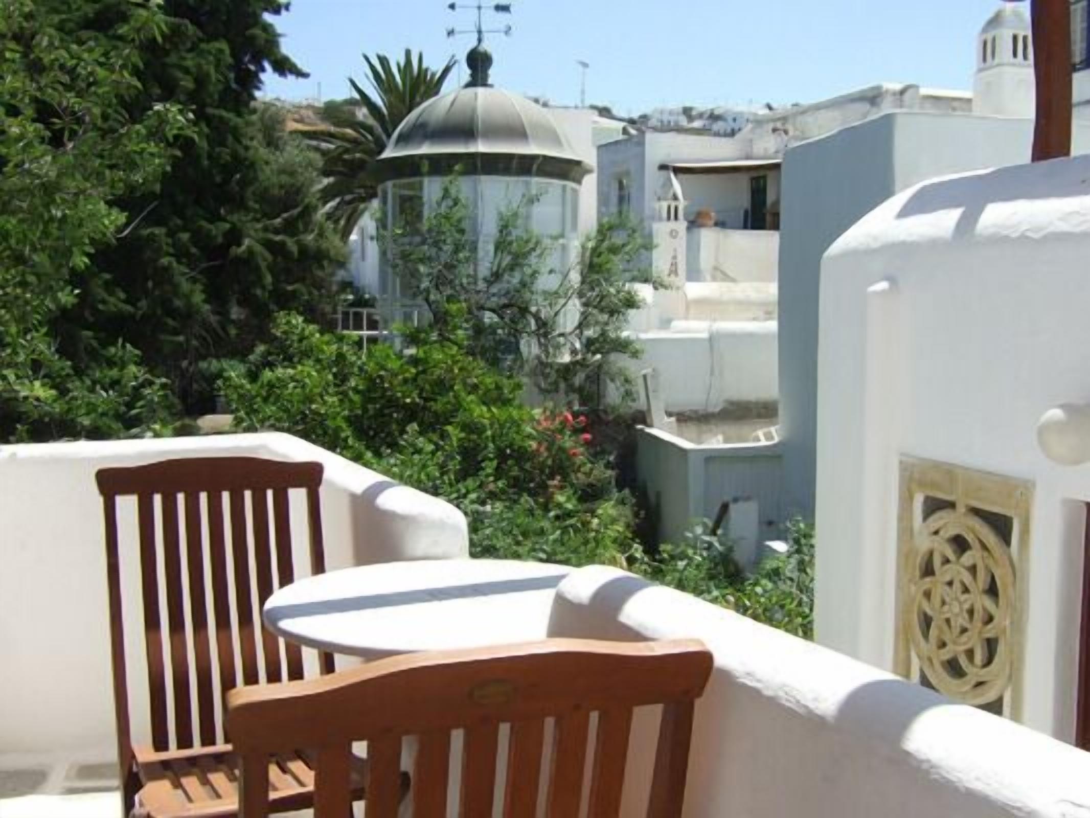 A balcony with a wooden chair and table, surrounded by greenery and buildings with white and blue facades.