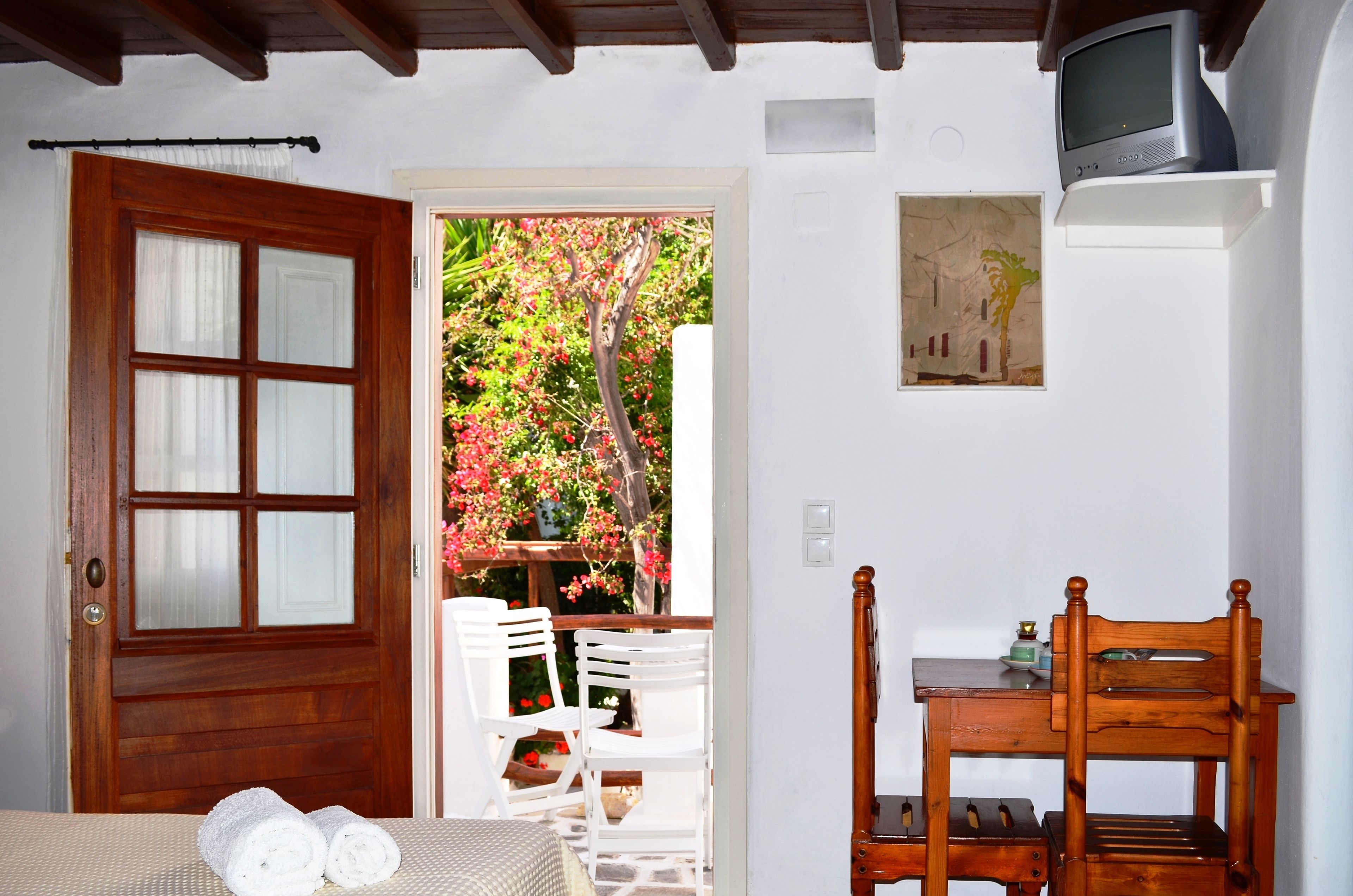 Una stanza con una porta in legno, un tavolo bianco con sedie, una sedia di legno, un tavolino con una radio e un quadro appeso al muro.
