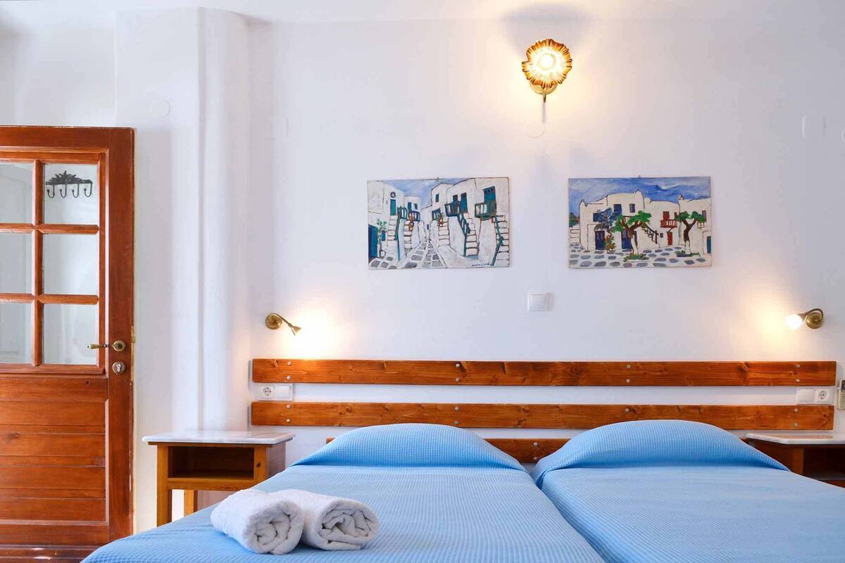Ein Hotelzimmer mit einem hölzernen Kopfteil, zwei Betten mit blauer Bettwäsche, einer Holztür und zwei an der Wand befestigten Bildern von weißen Gebäuden.
