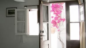 A room with a bed, a door leading outside, and a window with pink flowers. - Villa Pinelopi (Mykonos)