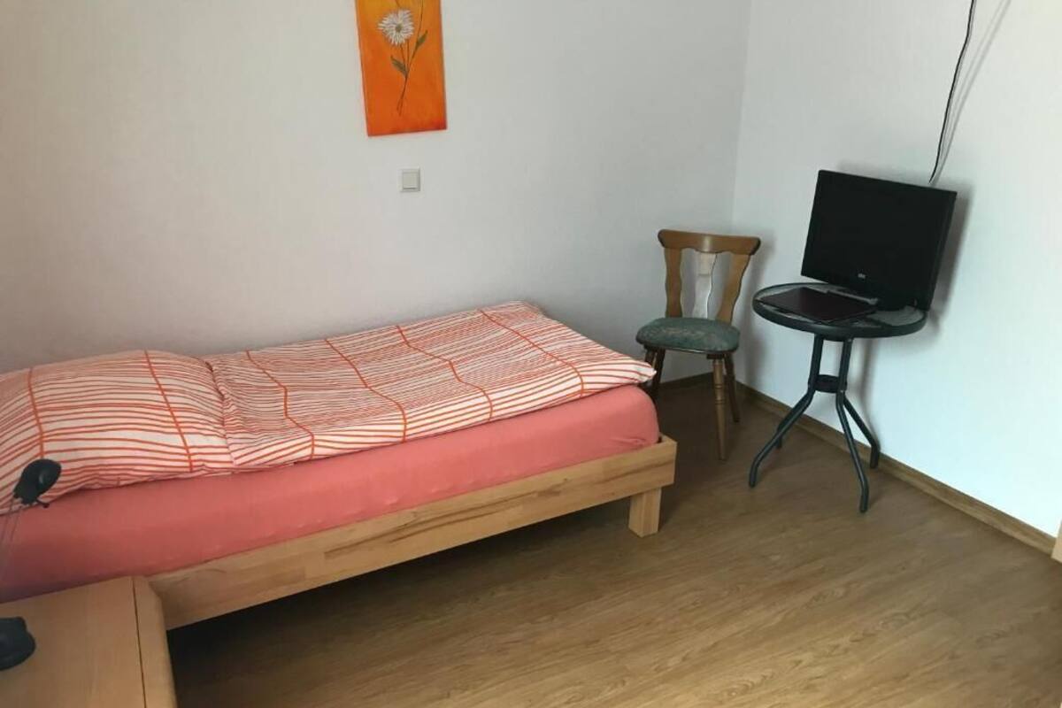 A small room with a single bed, a chair, a table with a laptop, and a TV.