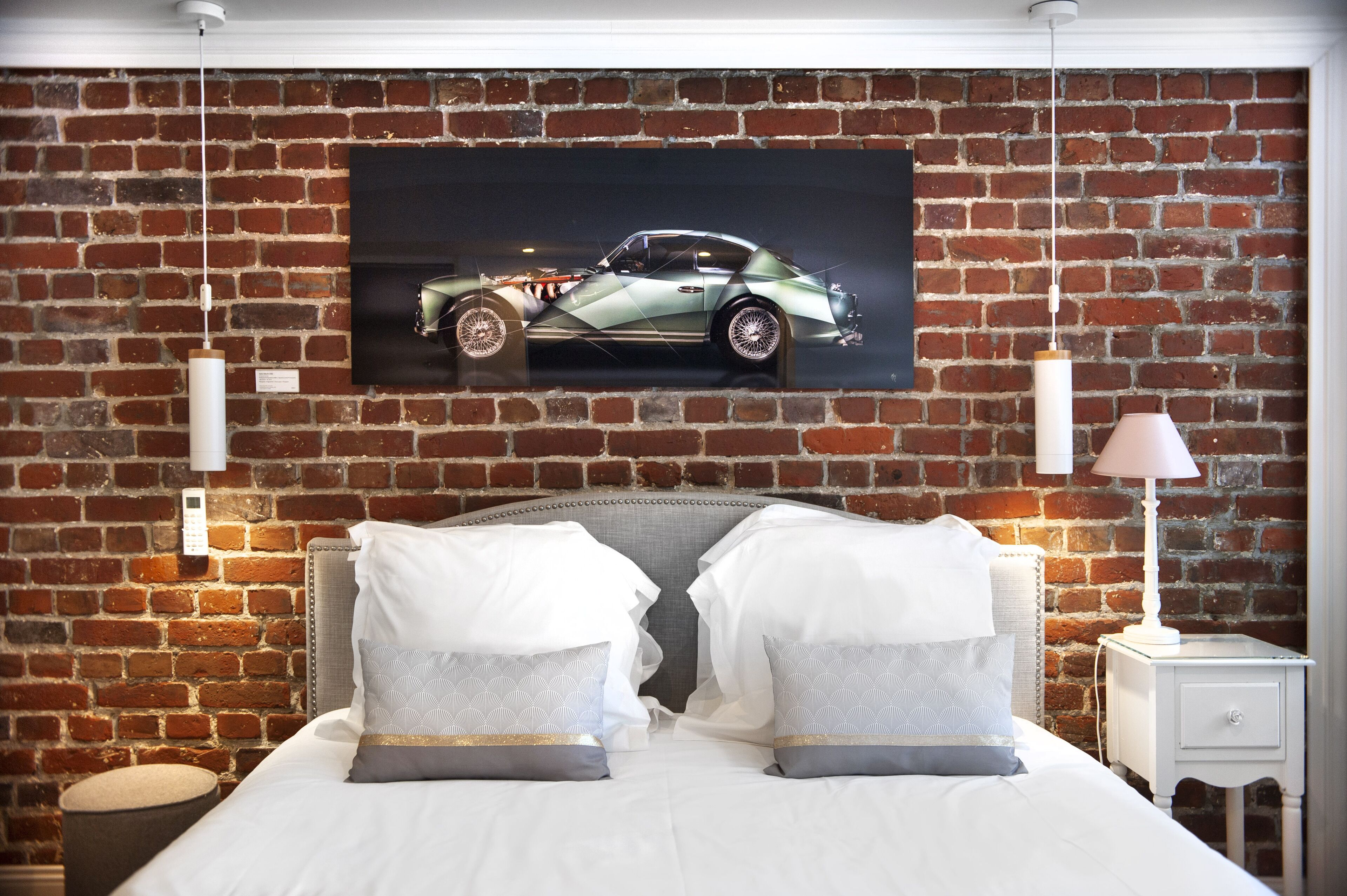 Une chambre d’hôtel avec un mur en briques, un lit avec une literie blanche et grise, une table de chevet avec une lampe et, au mur, un tableau représentant une voiture.