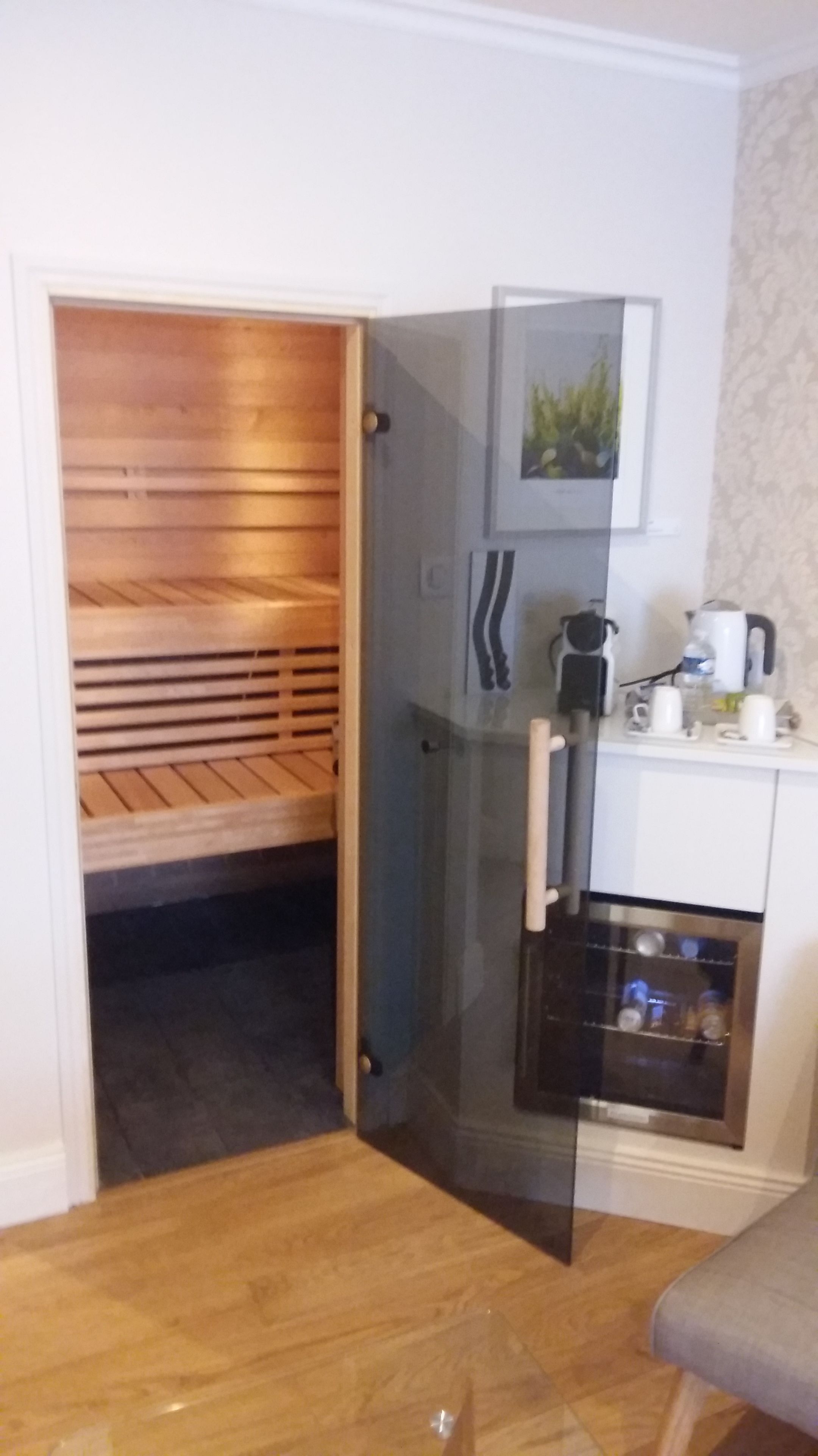 Ett rum med bastu, en glasdörr som leder till ett köksområde med vattenkokare och koppar, och en träbänk.