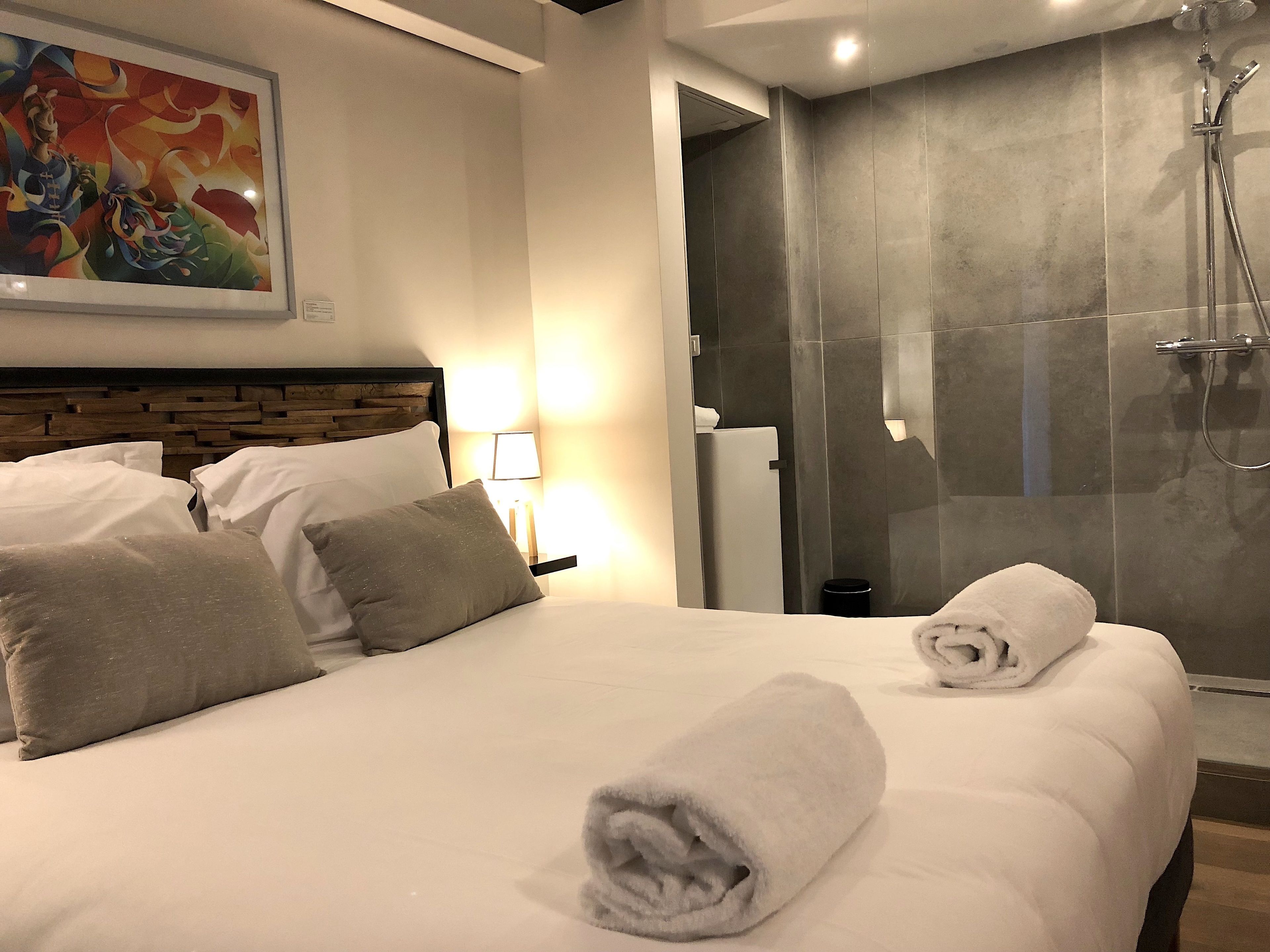 A hotel room with a large bed, a nightstand with a lamp, a shower area, and a colorful abstract painting on the wall.