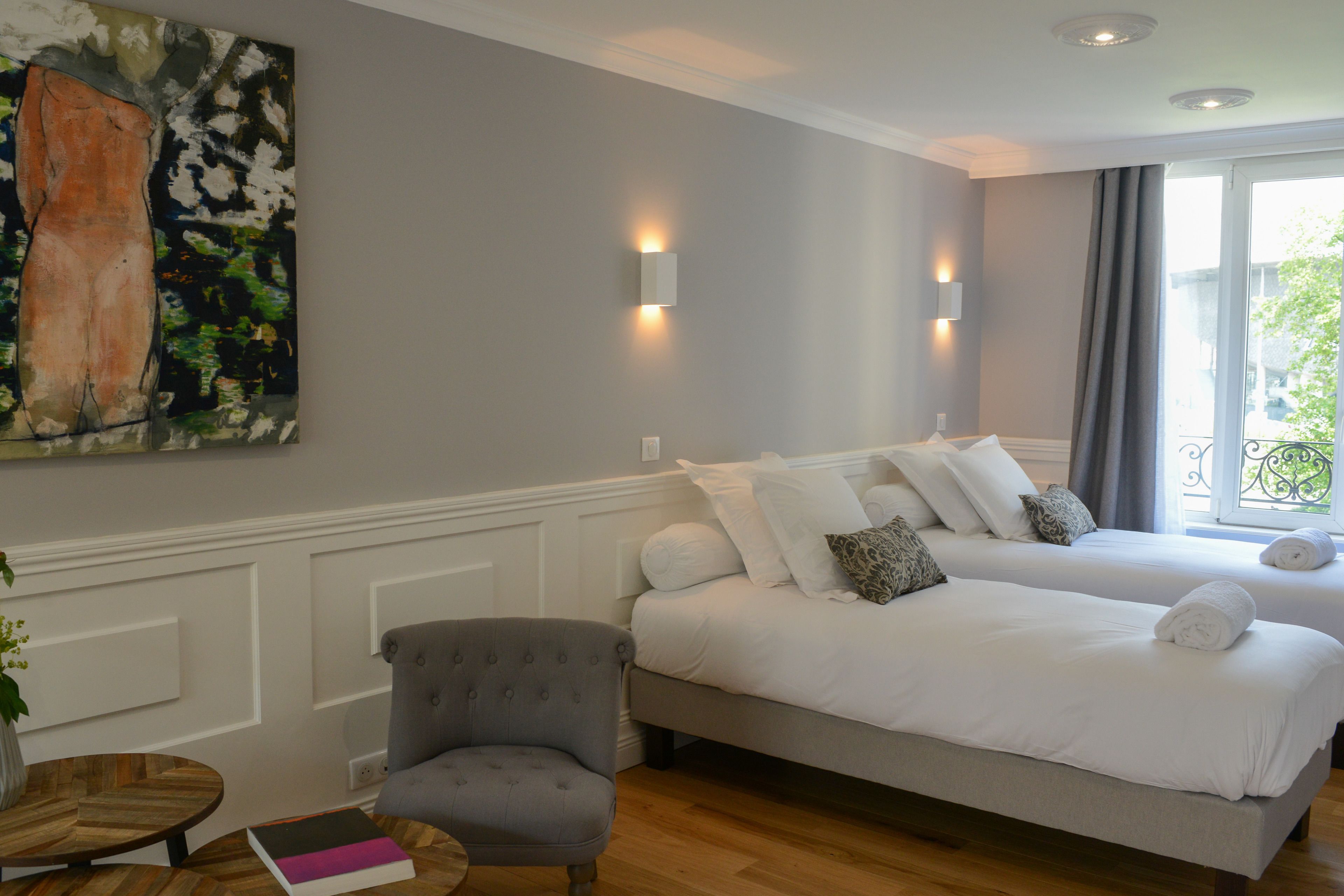 A hotel room with two beds, a grey armchair, a wooden side table, and a painting on the wall.