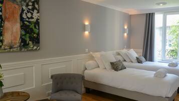 A hotel room with two beds, a grey armchair, a wooden side table, and a painting on the wall.