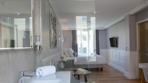 Una habitación de hotel moderna con un espejo grande, un lavamanos blanco, una cama con ropa de cama blanca, una pequeña mesa redonda con un jarrón de flores y un televisor montado en la pared.