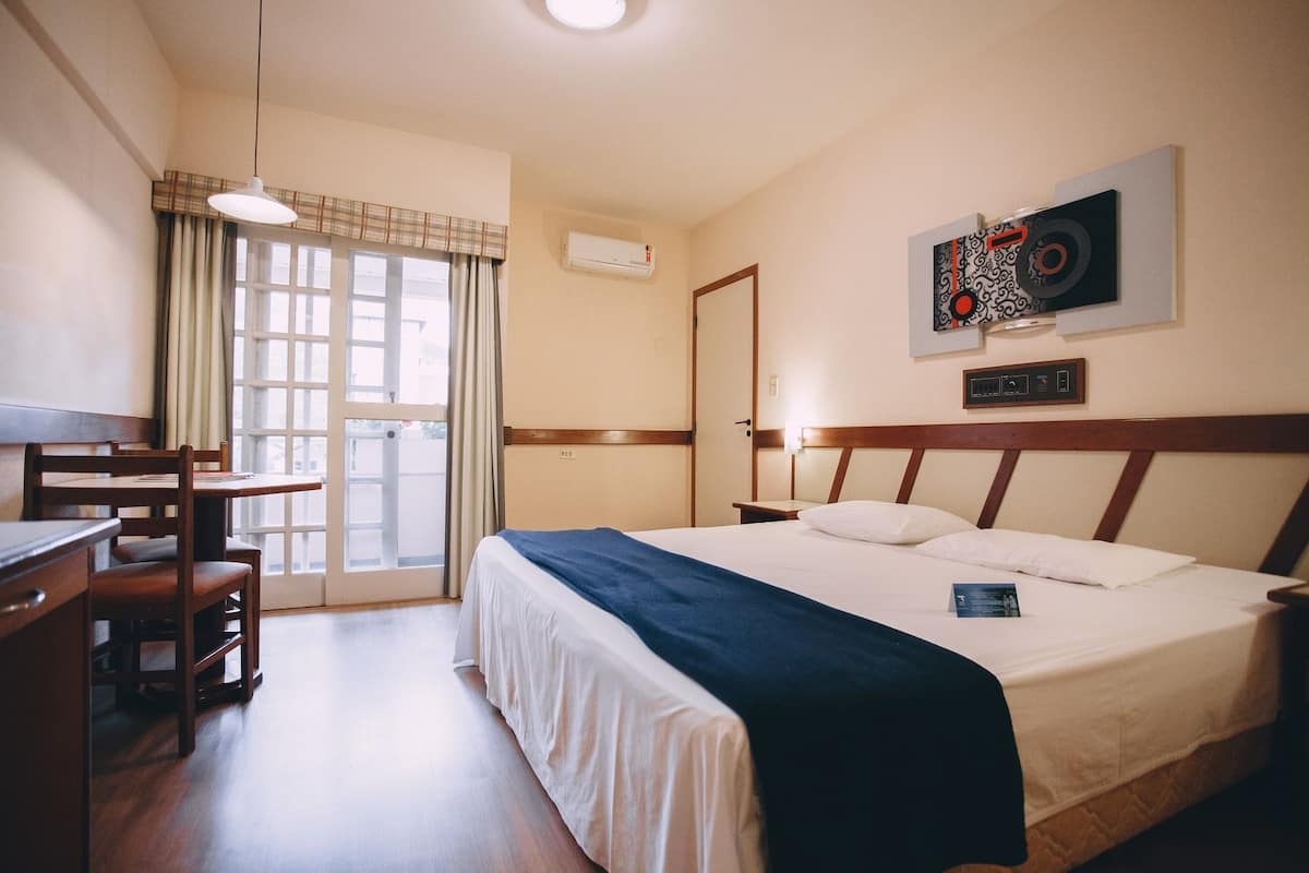 Ein Hotelzimmer mit Bett, Schreibtisch und Stuhl. Es gibt ein Fenster mit Vorhängen, eine Wand-Klimaanlage und eine Wanddekoration.
