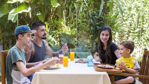 Uma família jantando ao ar livre em uma mesa com toalha branca, rodeada por vegetação exuberante.