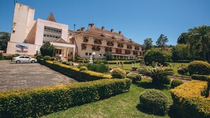 Um hotel com um edifício moderno e um edifício tradicional com varandas. Há um carro estacionado na frente e um jardim bem cuidado, com sebes e árvores.