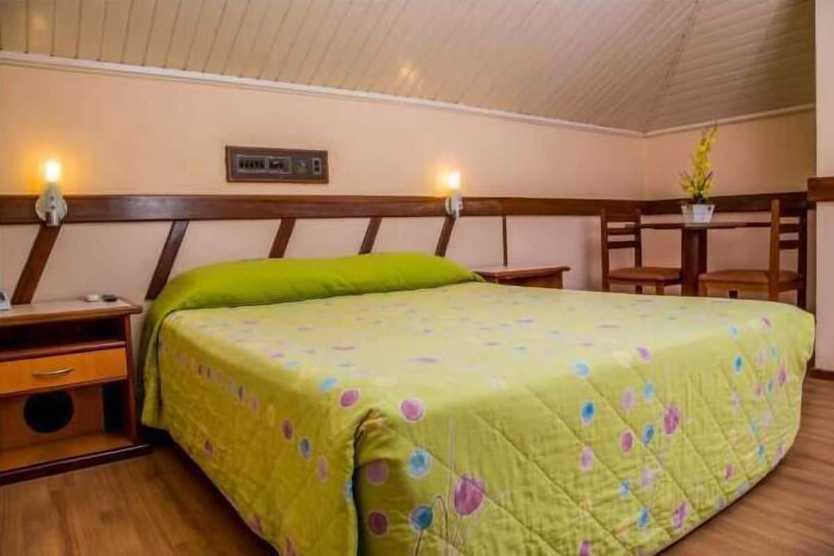 Ein Zimmer mit einem Einzelbett, einer grünen Tagesdecke, einem Holz-Kopfstück, Nachttischen und einem kleinen Tisch mit Stühlen.