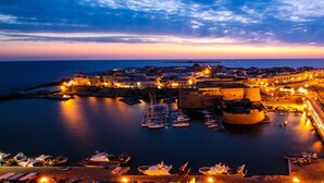 Une ville côtière au crépuscule, avec un port, des bateaux et des bâtiments illuminés.