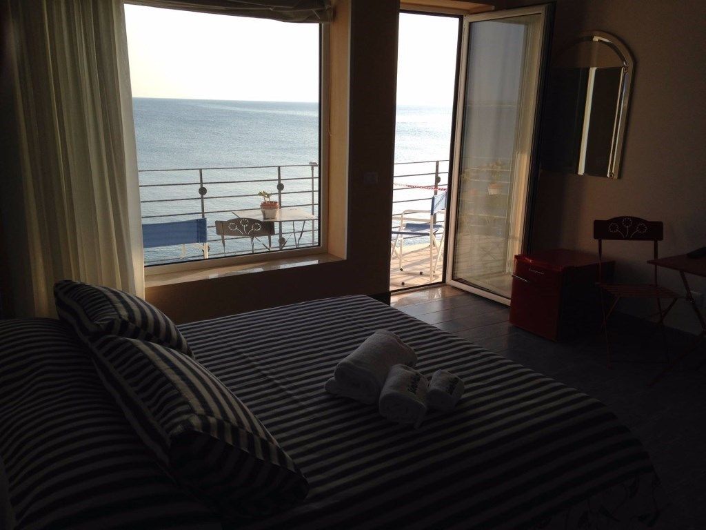 Une chambre d’hôtel avec un lit, une fenêtre donnant sur la mer, un balcon avec des chaises et une petite table.