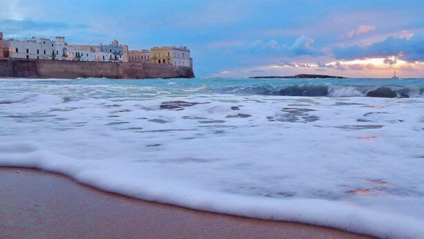 Un paysage côtier avec un ensemble de bâtiments au bord de la mer, des vagues qui s’écrasent sur le rivage et un coucher de soleil en arrière-plan.