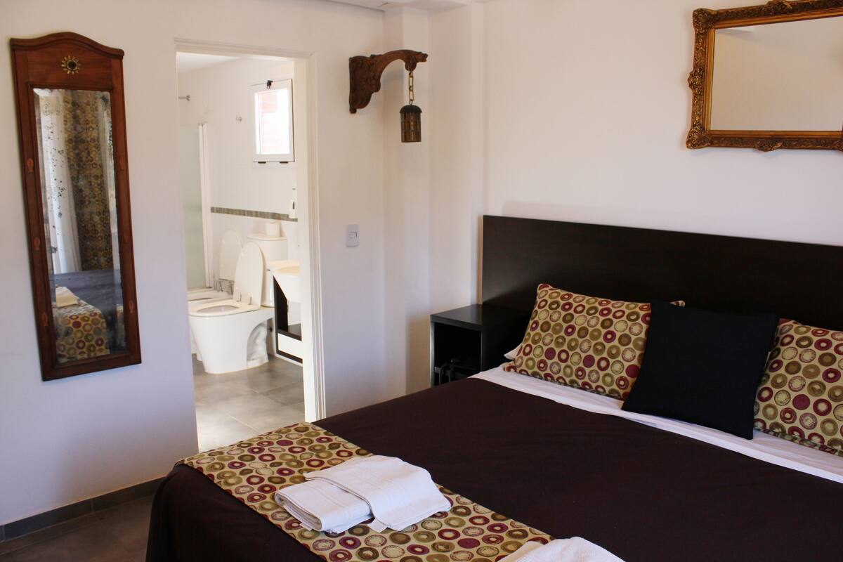 Ein Hotelzimmer mit einem großen Bett, einem Nachttisch, einem Spiegel und einem durch eine geöffnete Tür einsehbaren Badezimmer.