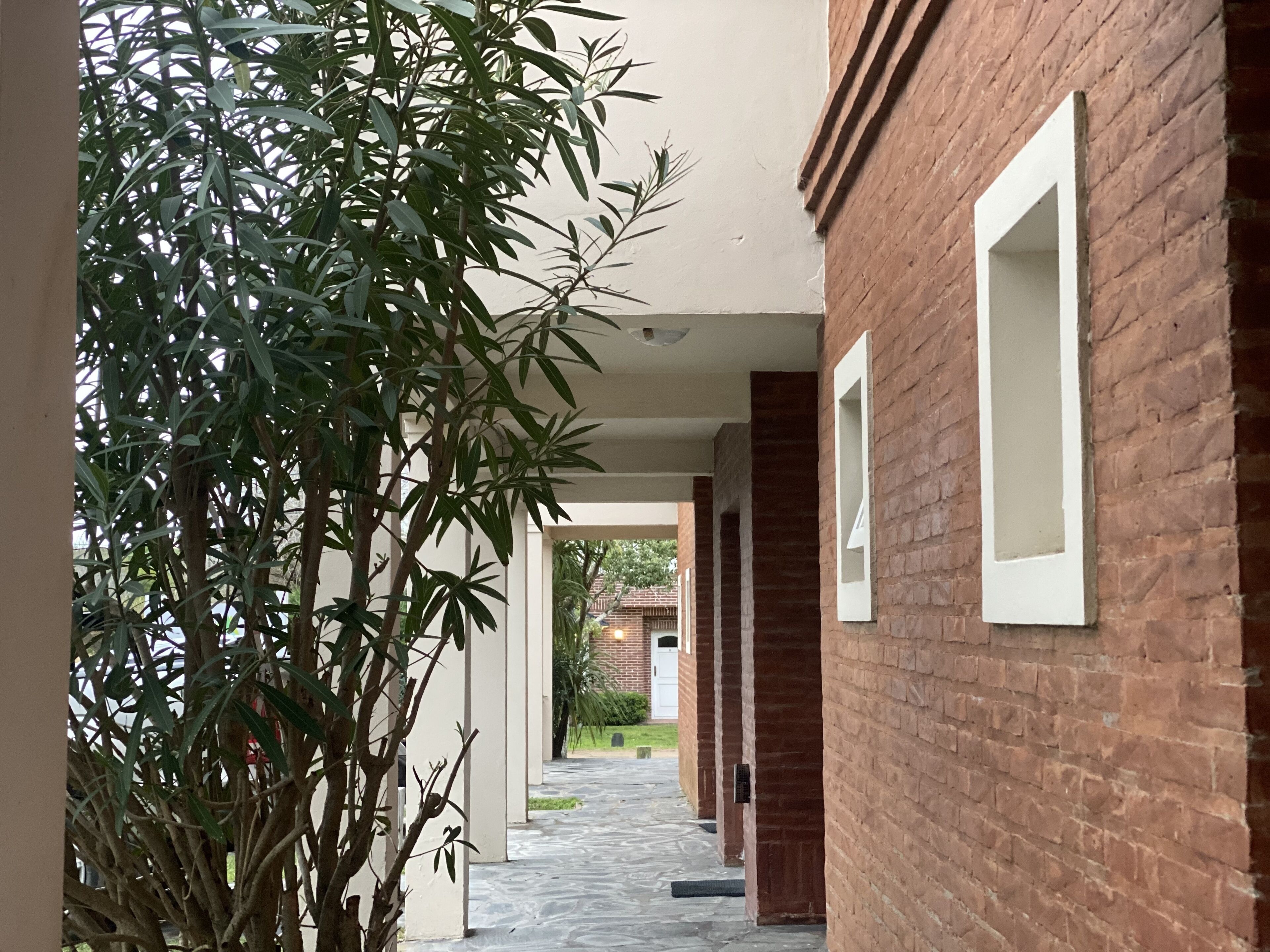A narrow brick corridor with white-framed windows, leading to a covered outdoor area.