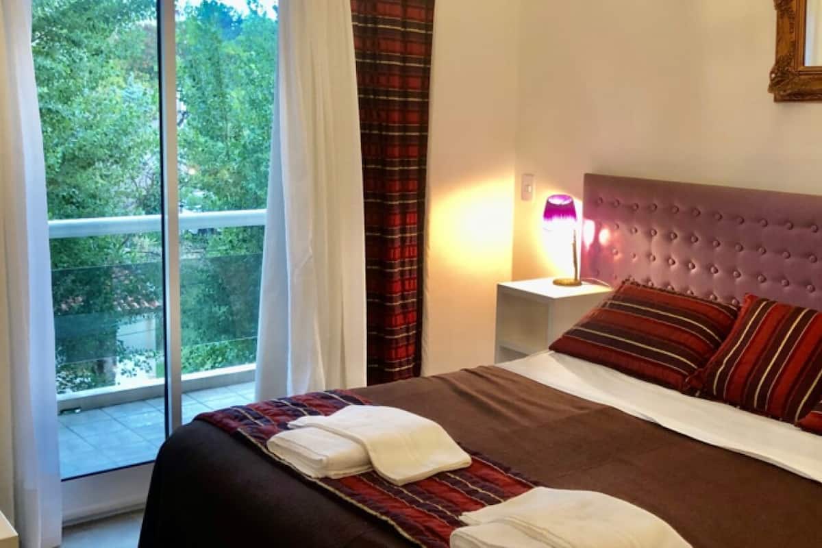 Ein Schlafzimmer mit großem Fenster, einem Bett mit gestreiften Kissen, einem gepolsterten Kopfteil und einem Nachttisch mit Lampe.