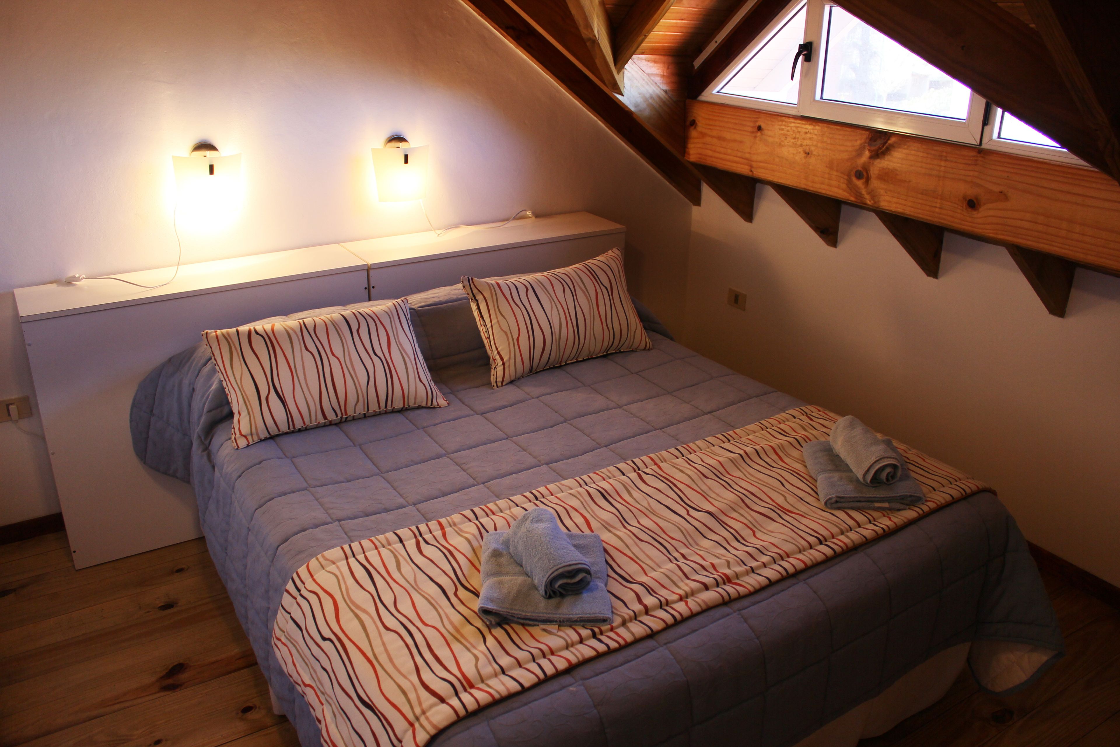 A bedroom with a sloped ceiling, a bed with striped bedding, two bedside lamps, and a window above the bed.