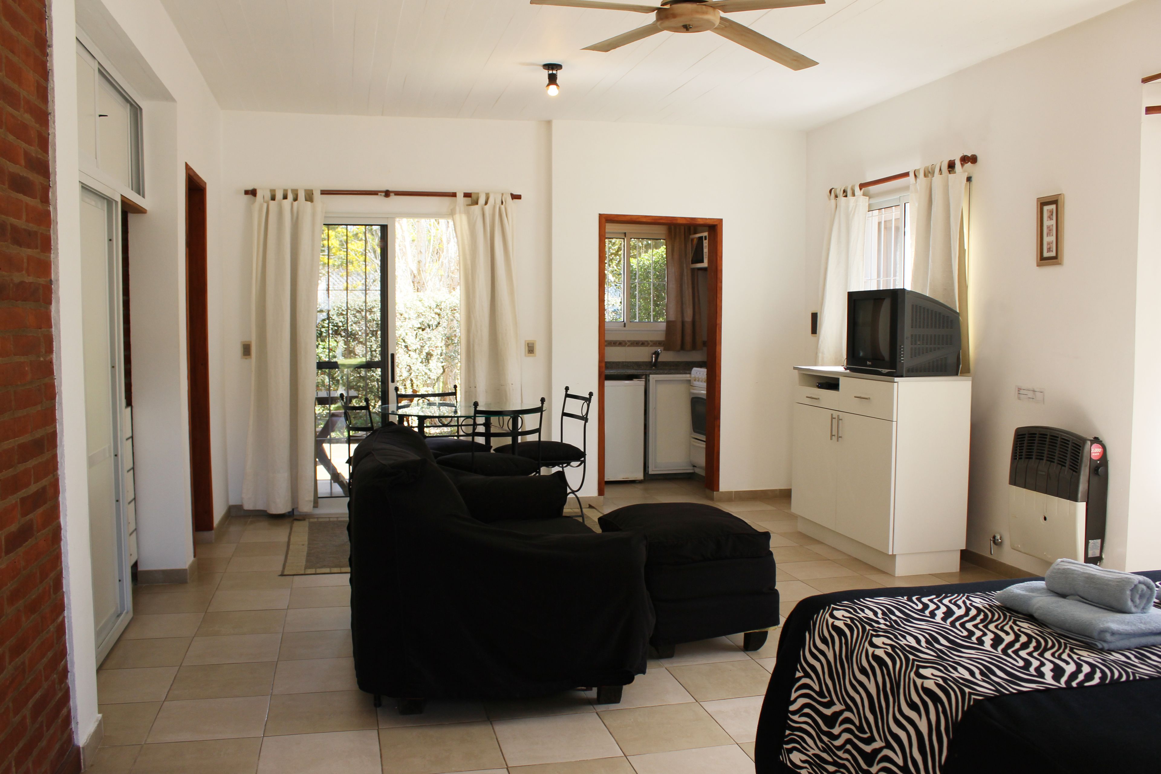 Una habitación con una cama, un sofá negro, una mesa de comedor con sillas, una zona de cocina y un balcón con cortinas.