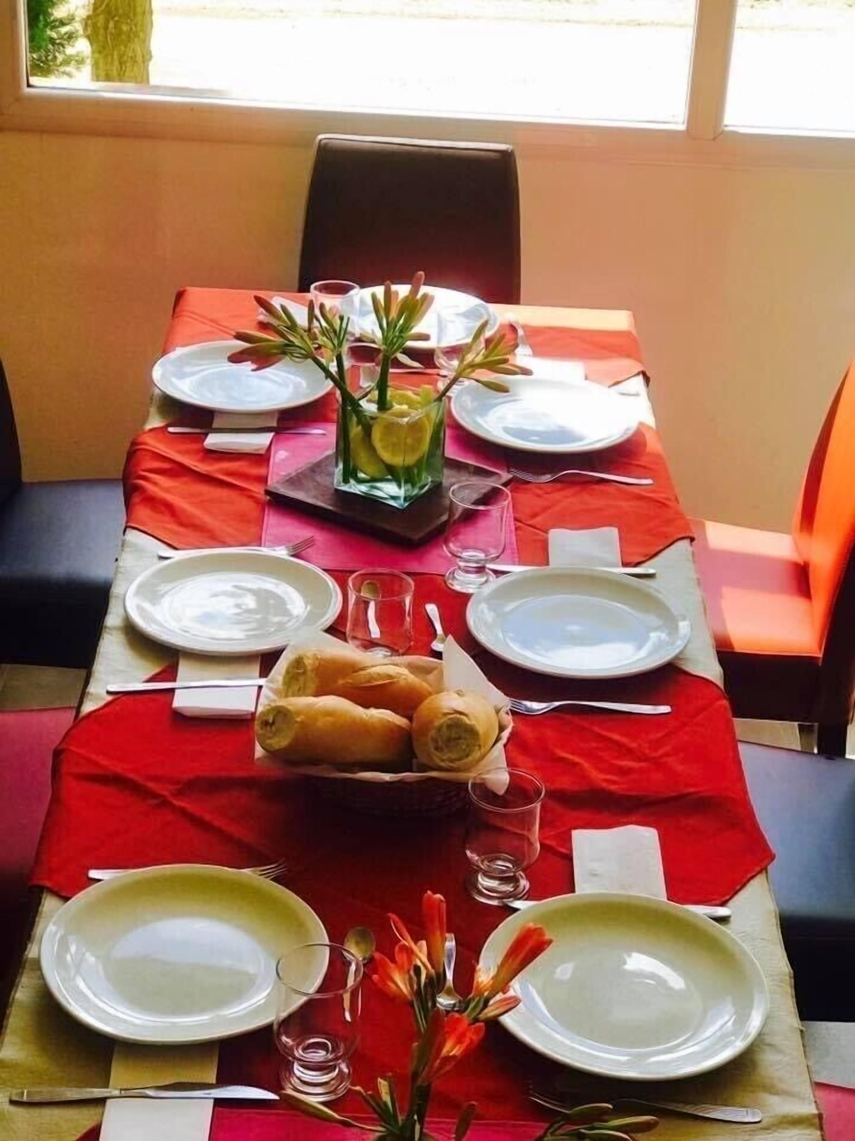 A dining table set for four with plates, cutlery, and a centerpiece of bread rolls and a glass vase with flowers.