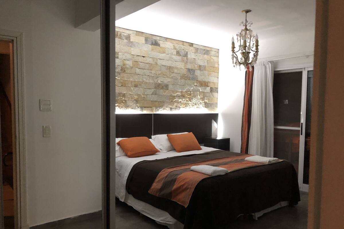 Ein modernes Schlafzimmer mit einer Wand aus Stein, einem großen Bett mit orange-weißer Bettwäsche, einer Chandelier und einer Schiebeglastür zu einem Balkon.
