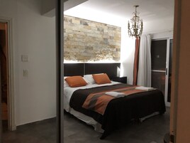 Un dormitorio moderno con una pared de piedra decorativa, una cama grande con ropa de cama naranja y blanca, un candelabro y una puerta corrediza de cristal a un balcón.